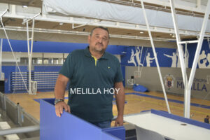 Man in a teal polo leaning on a blue railing inside an indoor basketball gym; blue walls and bleachers behind him, with the text 'MELILLA HOY' overlaid.