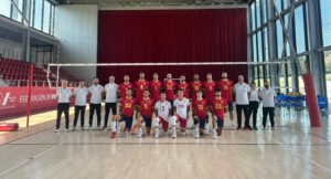 Group photo of a men's volleyball team in red and yellow uniforms posing with coaches on an indoor court near the net.