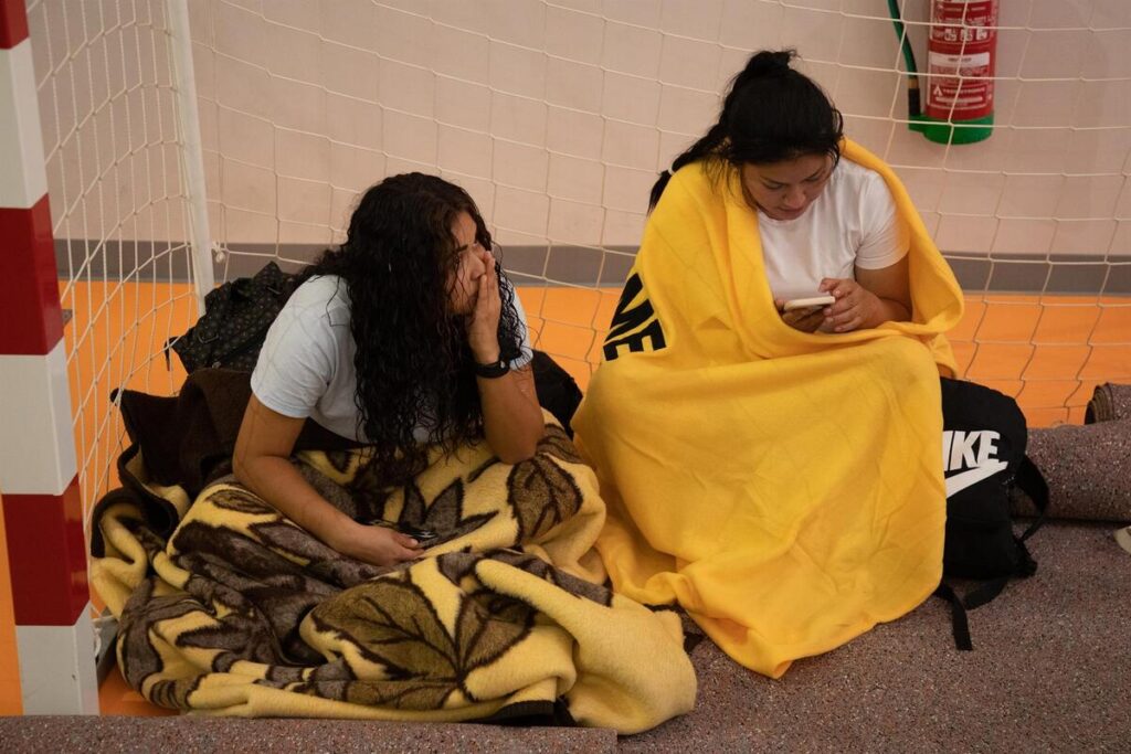 Two friends sit on an orange gym floor wrapped in blankets, one scrolling on a phone.