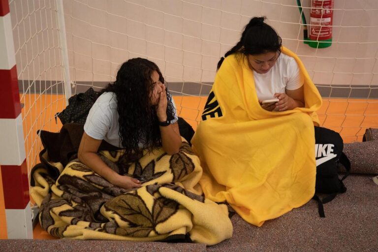 Two friends sit on an orange gym floor wrapped in blankets, one scrolling on a phone.