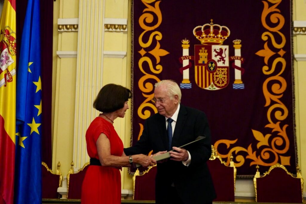 Two formally dressed individuals shake hands on a stage as a certificate is exchanged, with Spain and EU flags and a regal purple banner in the background.