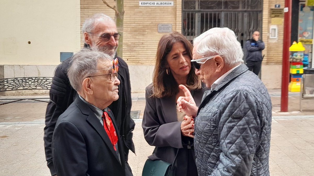 Four adults standing on a city sidewalk engaged in conversation, the man with white hair and sunglasses gesturing with a finger in the air.