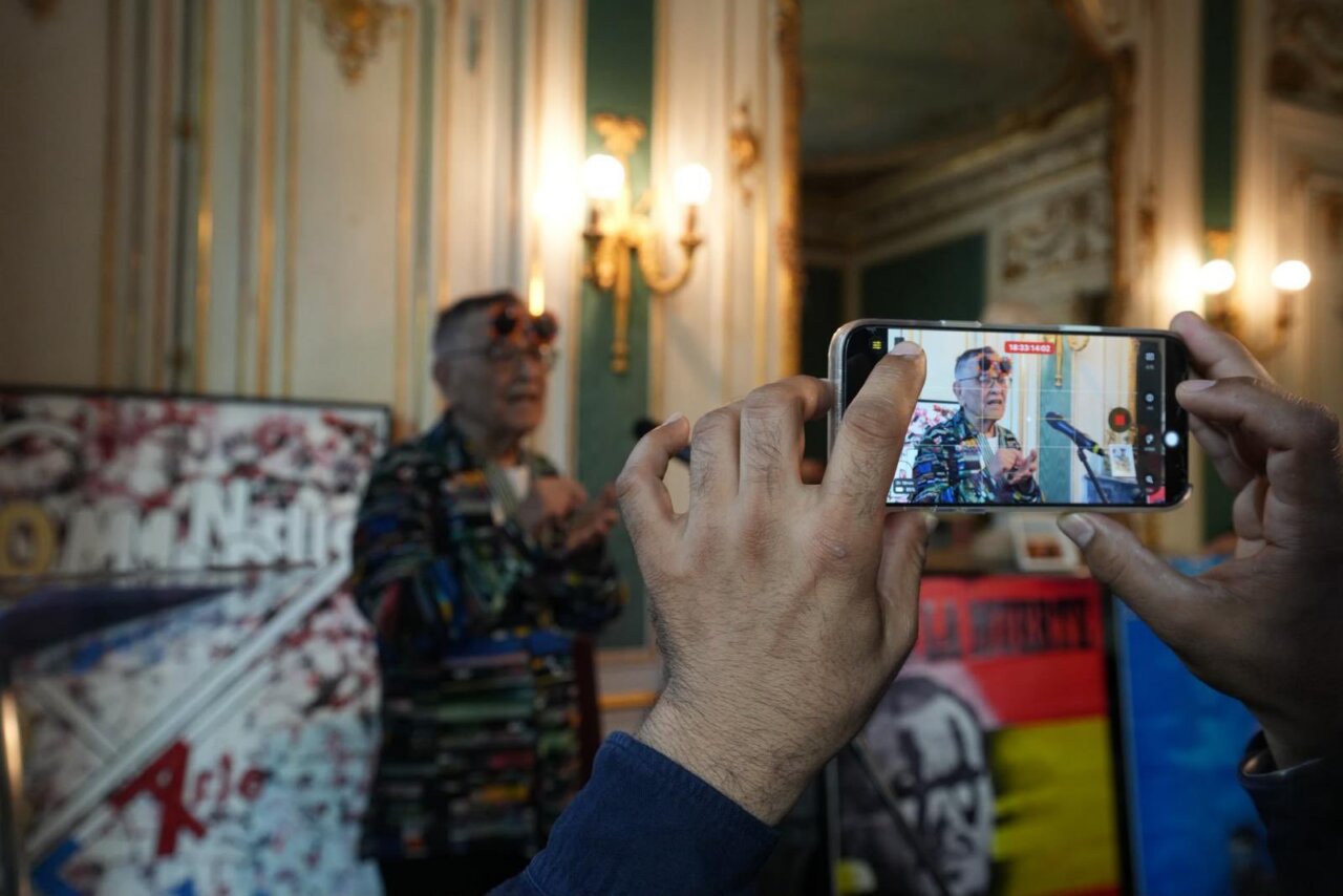 Person in a patterned shirt speaks at a press event in an ornate room, filmed with a smartphone held up in the foreground