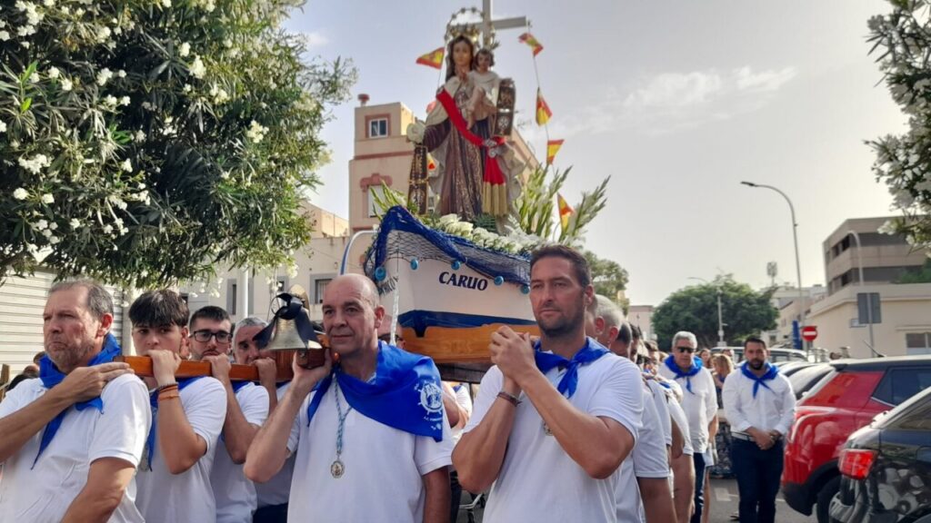 Group of men in white shirts and blue scarves carry a decorated float with a statue of the Virgin Mary through a street during a religious procession.