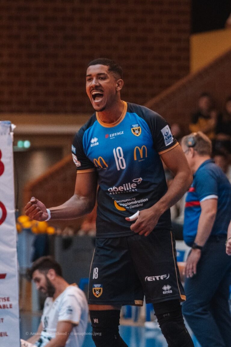 Male athlete in a blue #18 jersey celebrating on an indoor court, fist clenched and mouth open in shout.