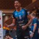 Male athlete in a blue #18 jersey celebrating on an indoor court, fist clenched and mouth open in shout.