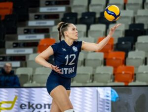 Female volleyball player in navy uniform with number 12 serves the ball on an indoor court, with colorful empty seats in the background and a decorative LED barrier nearby.