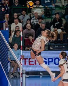 Female volleyball player in white leans back and spikes the ball mid-air near the net as spectators watch.