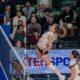 Female volleyball player in white leans back and spikes the ball mid-air near the net as spectators watch.