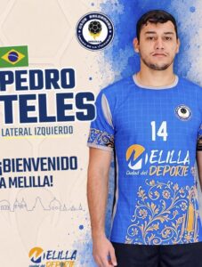 Male handball player in a blue jersey with the number 14, standing beside a welcome banner that says 'Bienvenido a Melilla' with the Melilla sports logo.