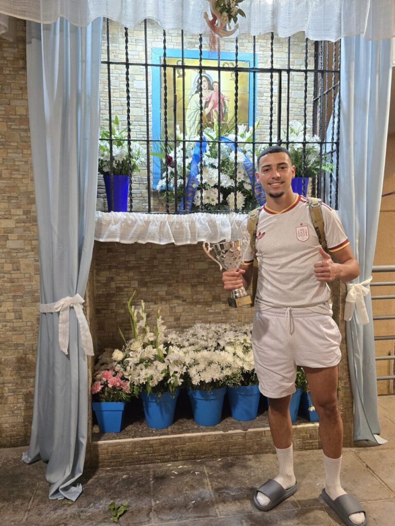 Man in white athletic outfit poses with a trophy in front of a flower-filled shrine featuring a framed religious image behind bars, giving a thumbs-up.