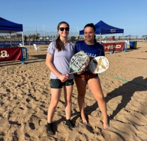 Two young women on a sunny beach posing with paddle tennis paddles in hand.
