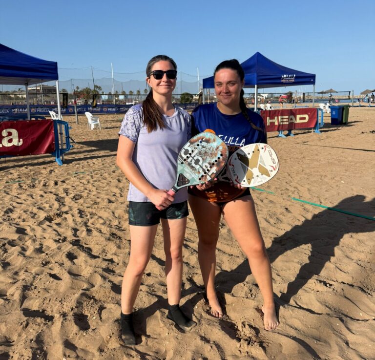 Two young women on a sunny beach posing with paddle tennis paddles in hand.