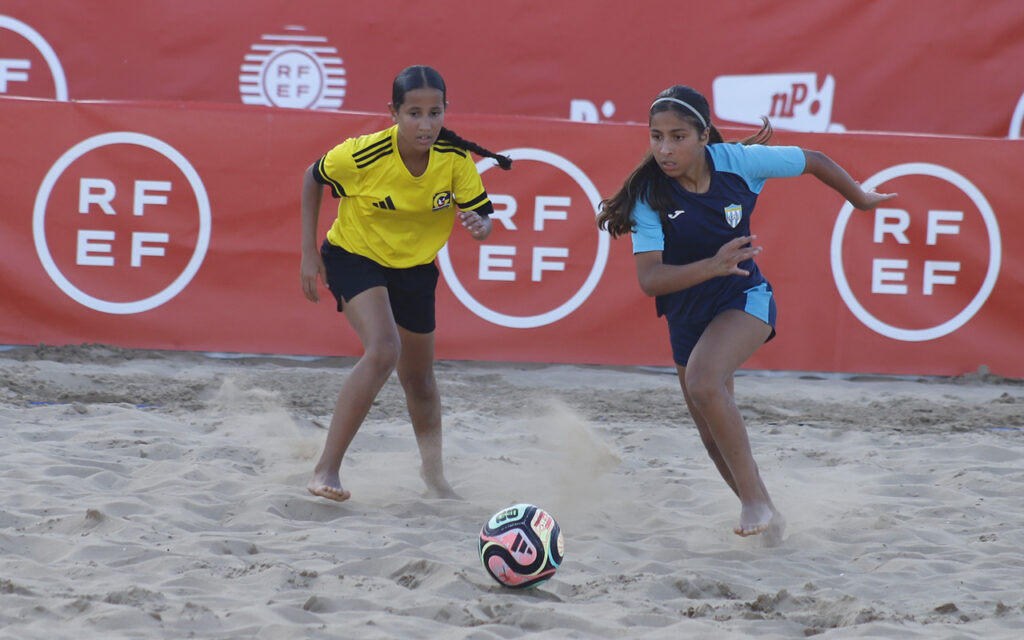 Two young girls playing beach soccer, one in a yellow jersey and black shorts, the other in blue, chasing a colorful ball as dust rises on the sand with red banners behind.