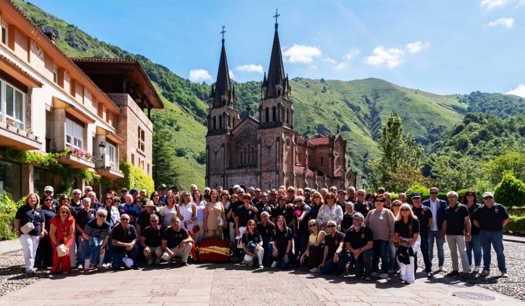 Large group of tourists posing for a photo in front of a stone church with twin spires and a green mountain backdrop.
