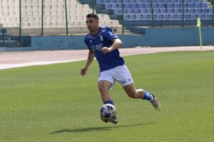 Soccer player in a blue jersey and white shorts dribbling a ball on a green field near empty stands.