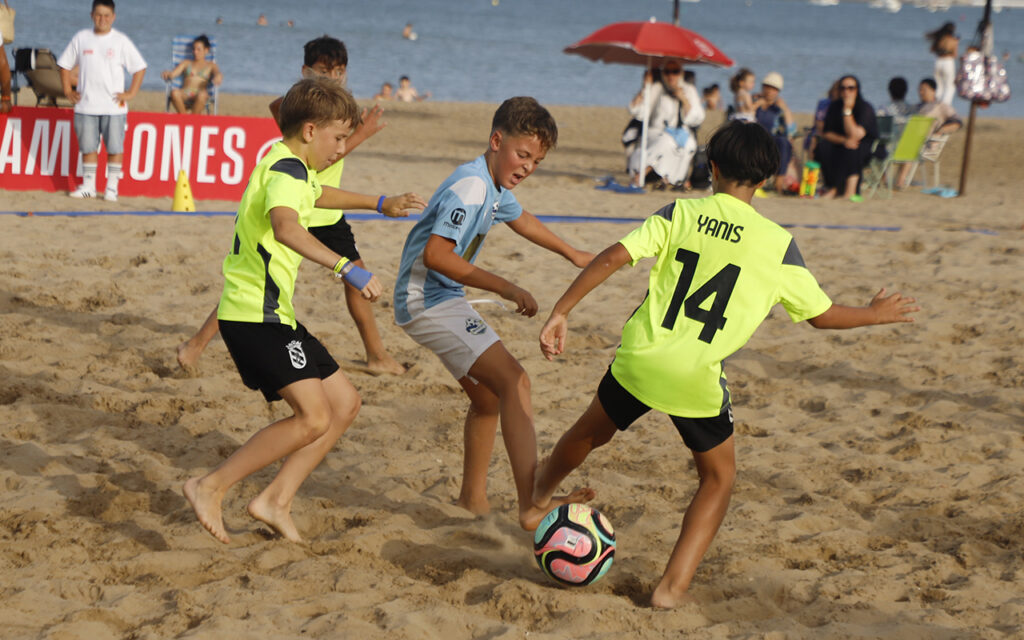 Young boys playing beach soccer on sand, chasing a colorful ball near spectators by the water.