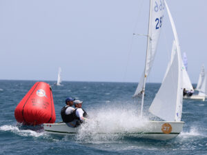 Two sailors in a small sailboat tack past a large red buoy on blue waters during a race, spray flying from the bow.
