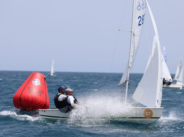 Two sailors in a small sailboat tack past a large red buoy on blue waters during a race, spray flying from the bow.