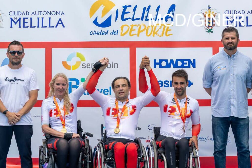 Three wheelchair athletes in white-red uniforms with gold medals lift their arms on a podium, flanked by two standing supporters; backdrop shows sponsor logos and 'Melilla' branding.