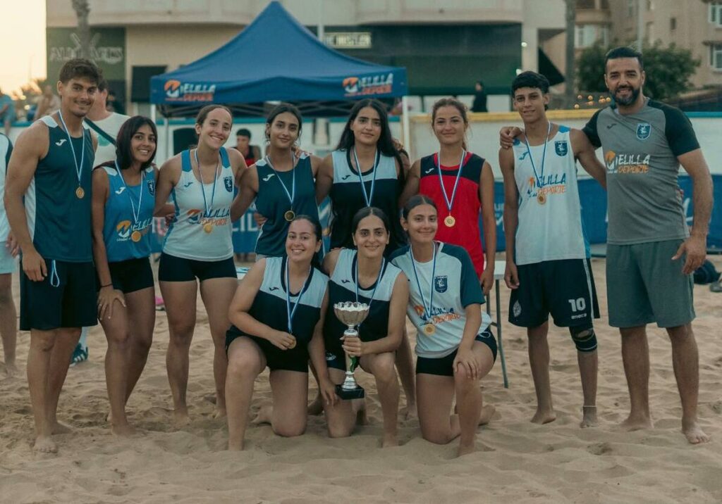 Beach volleyball team of 12 posing with medals and trophy after tournament, sand court and tents in the background.