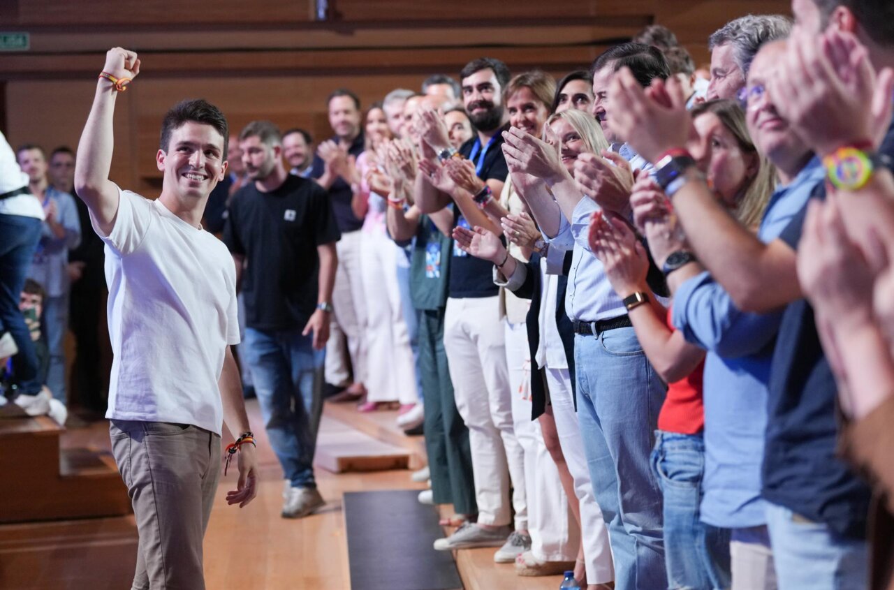 Smiling man in a white T‑shirt raises his fist in triumph as a crowd cheers and applauds behind him on stage or in a hall.