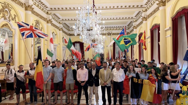 A diverse group of people in a grand gilded hall holding national flags, gathered under a crystal chandelier for a multicultural event (flags include UK, Brazil, Italy, Belgium).