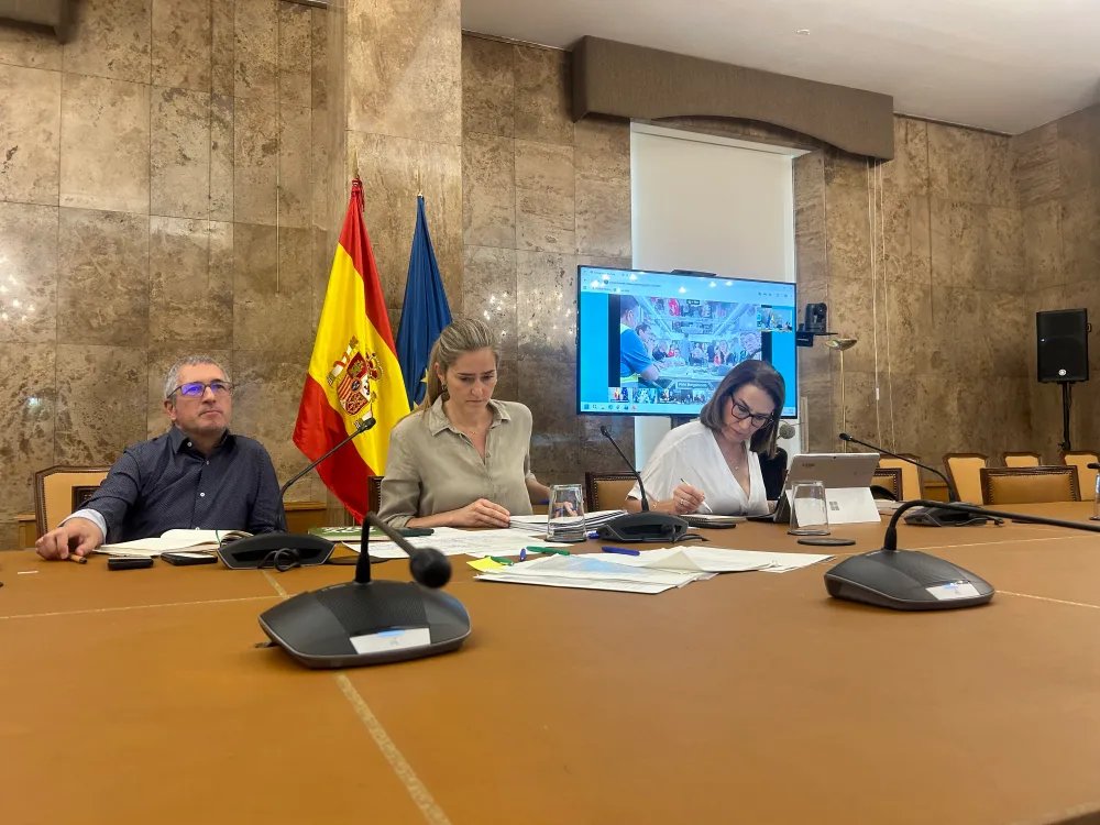 Reunión sobre incendios forestales en Madrid - MelillaHoy Three people sit at a long conference table in a marble-walled room, with Spain and EU flags behind them, microphones and documents in front of them.