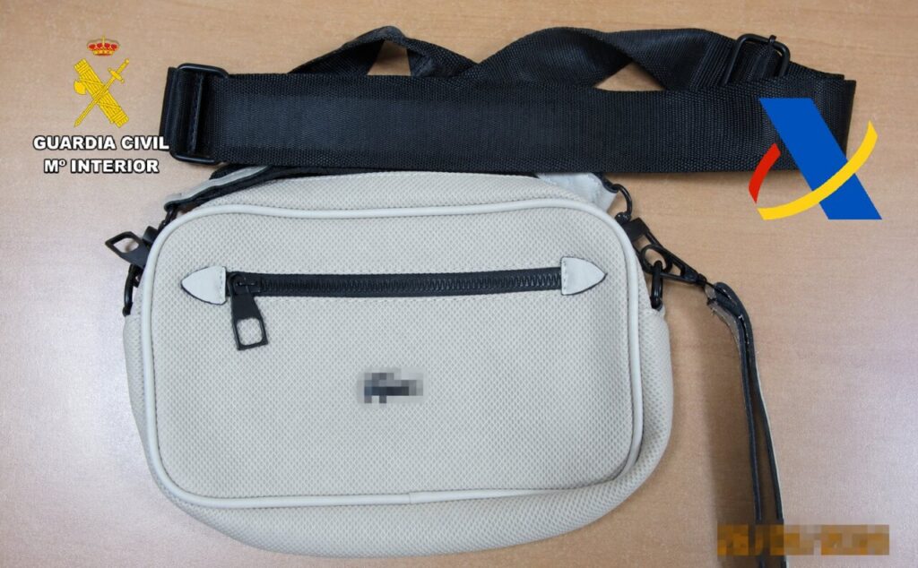 Beige small crossbody bag on a wooden table; left shows Guardia Civil emblem with 'GUARDIA CIVIL Mº INTERIOR', right has a blue-yellow-red logo.