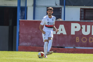 Soccer player in white uniform dribbling a ball on a grassy field near a red barrier backdrop.