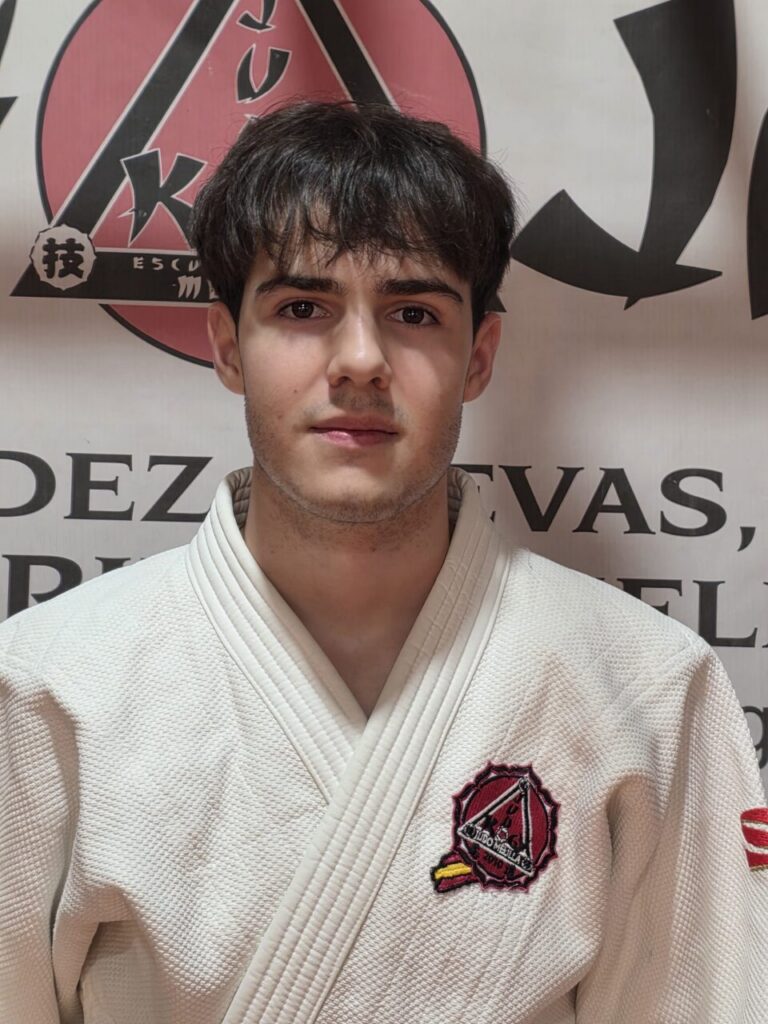 Young man wearing a white gi with a red emblem on the chest, standing in front of a wall with a martial arts logo behind him.