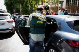 Guardia Civil officer with UCO vest opening the rear car door on a city street as reporters film nearby.