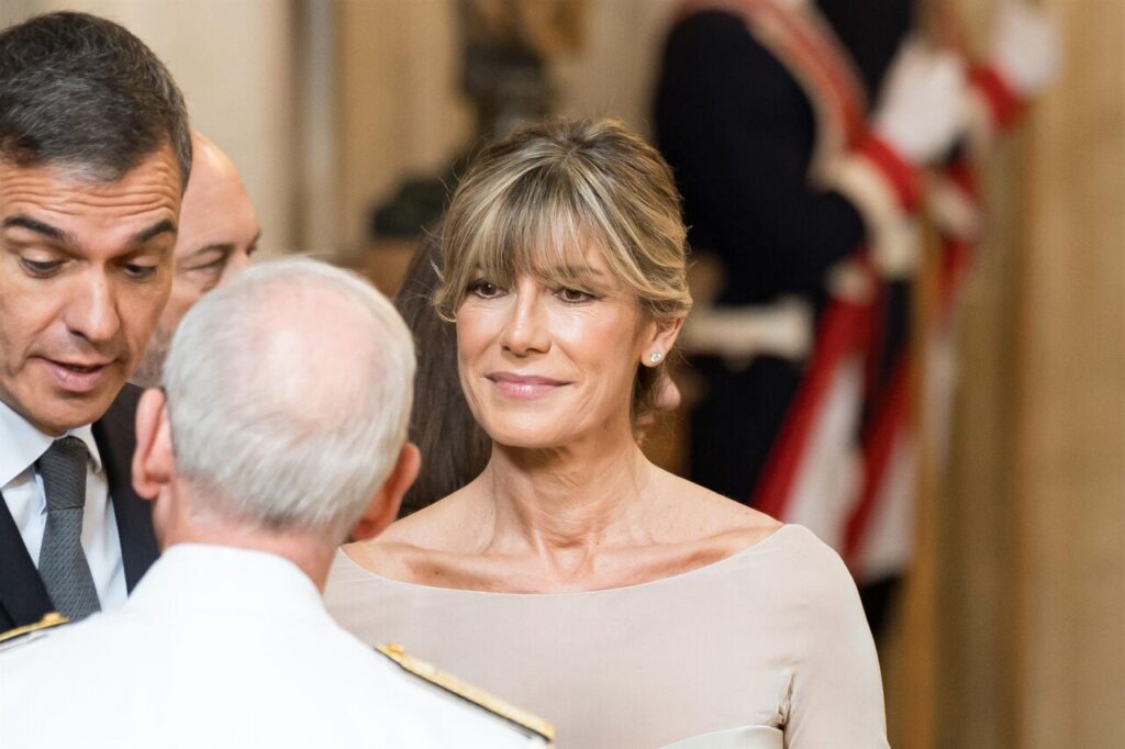 Woman in a beige dress smiles while talking with a man in a suit at a formal event; a uniformed person is in the foreground.