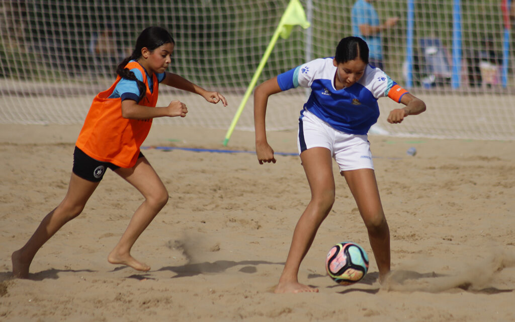 Two girls play beach soccer on sand; the girl in blue controls a multi-colored ball while the girl in orange pursues.