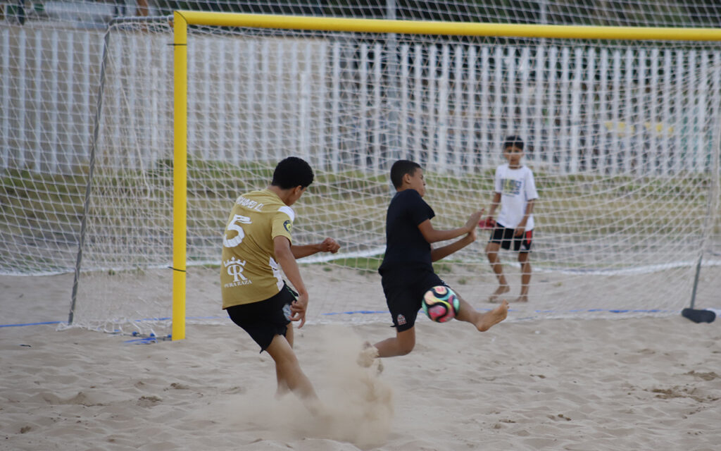 Two boys play beach soccer near a yellow goal; one in gold jersey kicks while the other in black attempts a ball kick, sand flying.