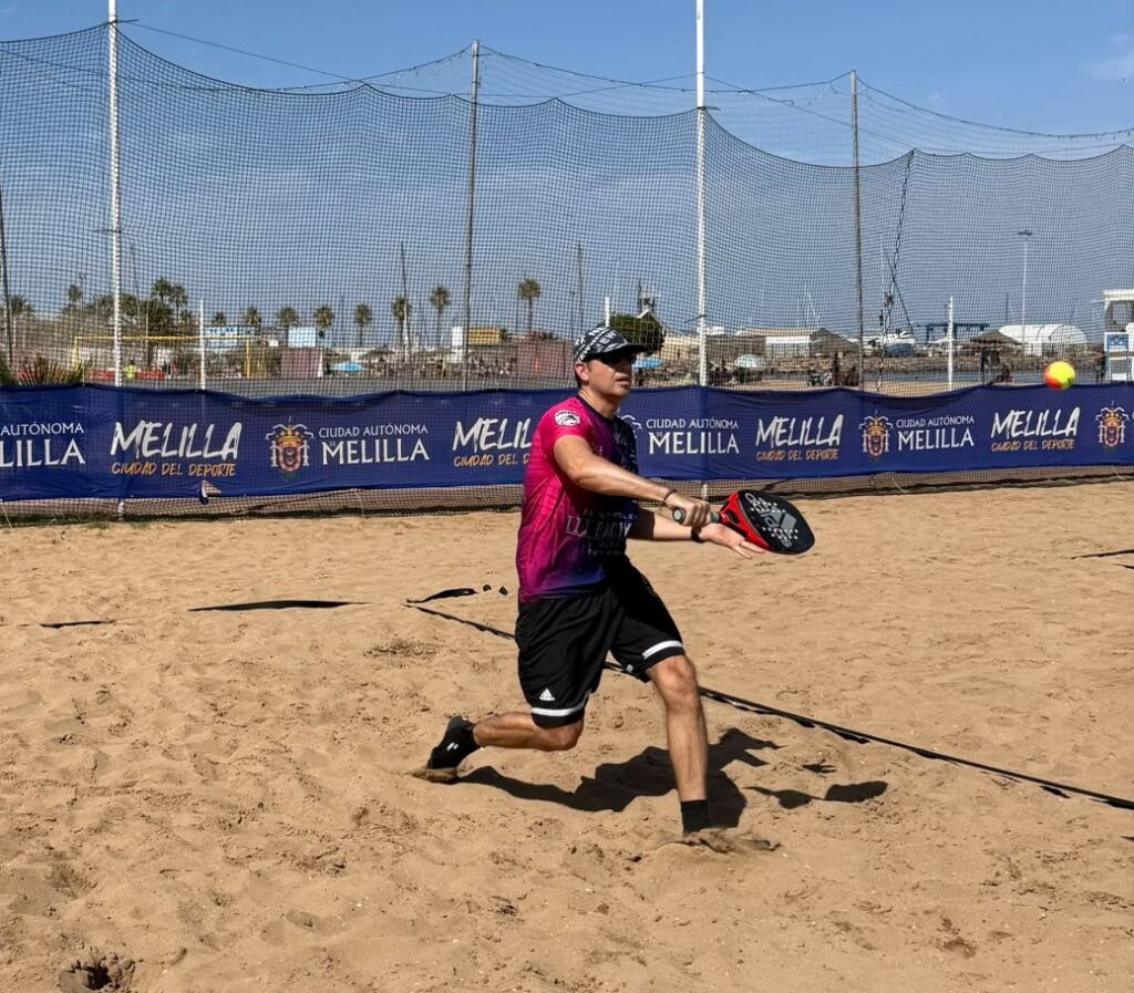 Male beach tennis player in a pink and black shirt swings a paddle on a sandy court with blue Melilla banners in the background.