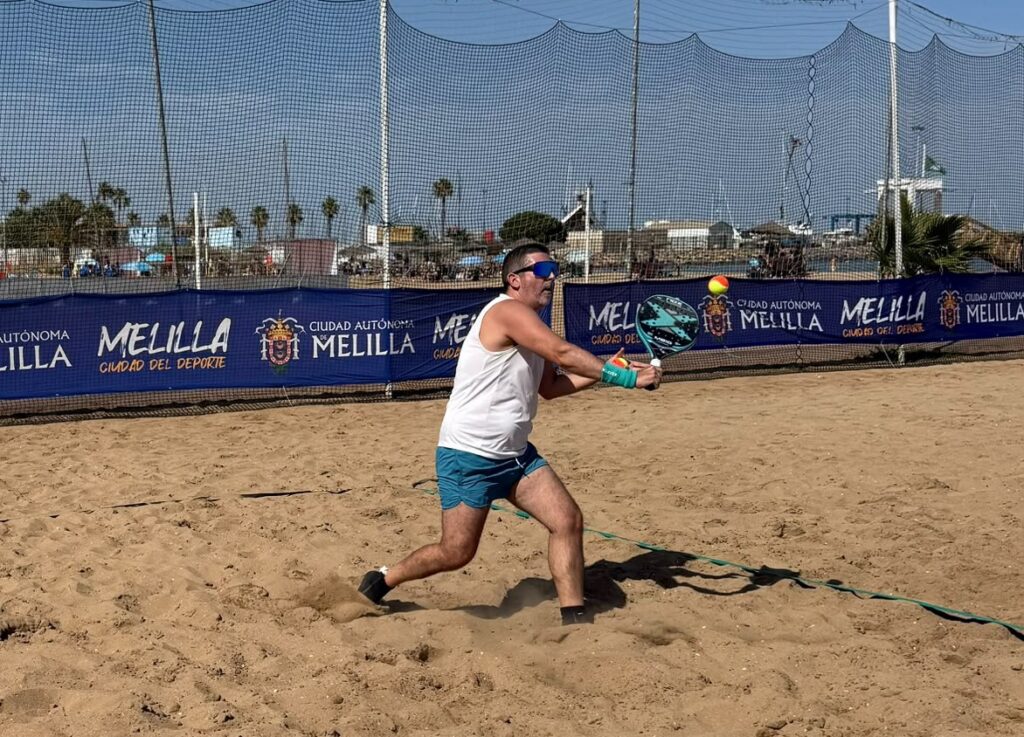 Man in a white tank top and teal shorts swings a paddle to hit a ball on a sandy beach tennis court, wearing sunglasses under a clear sky.