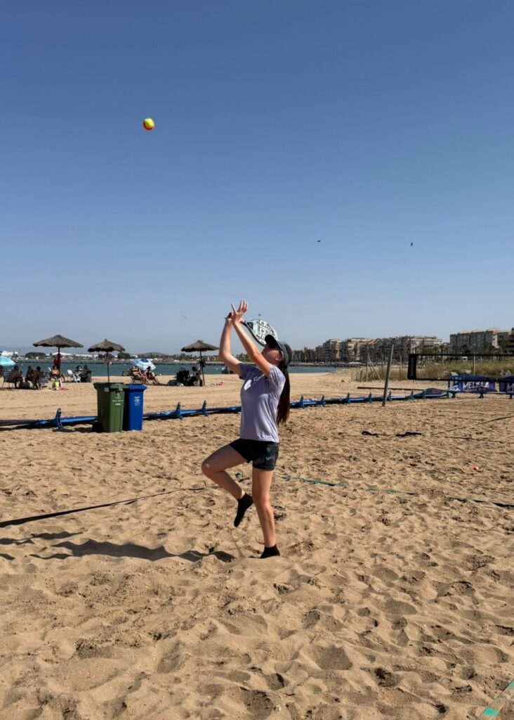 Woman on a sunny beach, balancing on one leg while throwing or catching a yellow ball in the air.