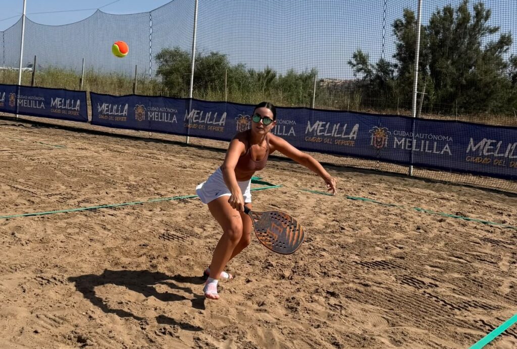 Woman playing beach paddle/tennis on a sandy court, mid-swing with a perforated paddle as a bright ball flies overhead.