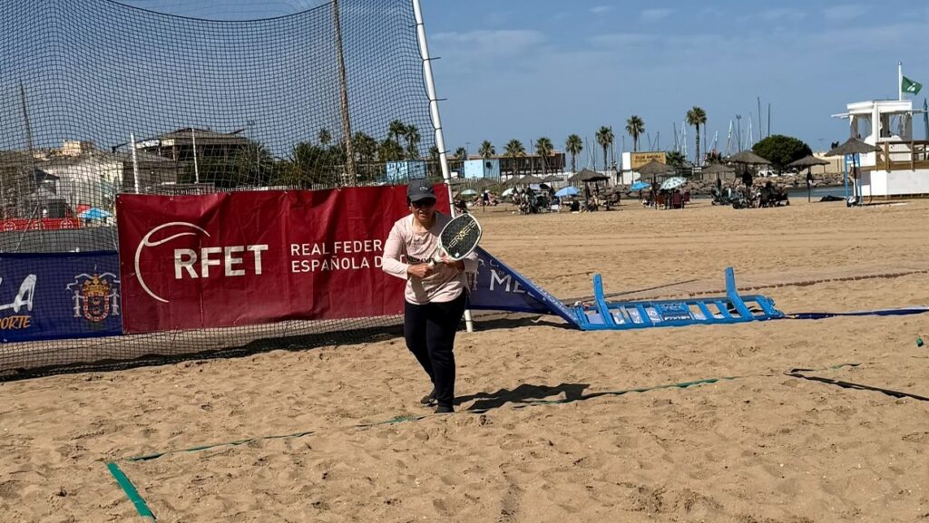 Person on a sunny beach holding a racket in front of a tennis practice net and red RFET banner, with spectators in the background.