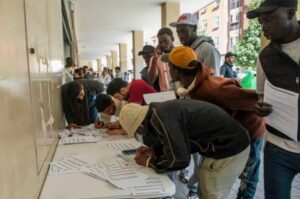 People line up at a long registration table under a covered walkway, signing and submitting forms outdoors at a busy event.