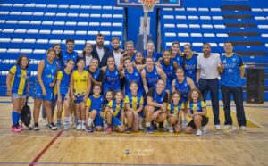 Group of basketball players and coaches posing for a team photo in a gym, wearing blue and yellow uniforms with a basketball hoop in the background