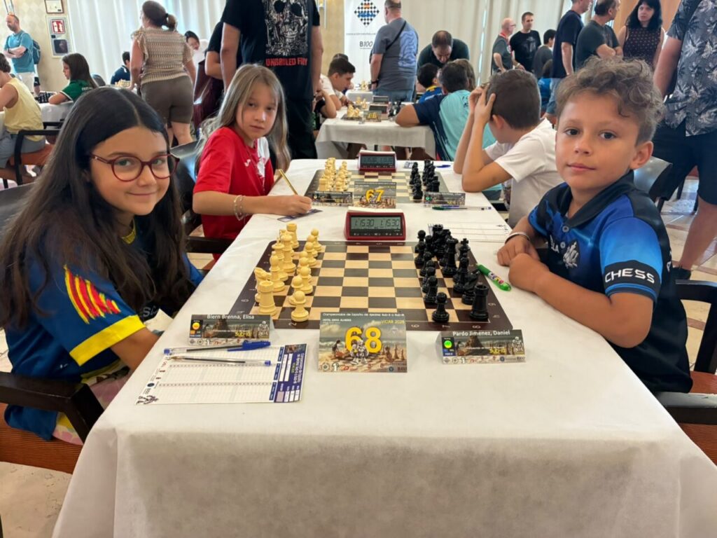 Three young chess players sit at a tournament table with boards, clocks, and score sheets, while spectators mingle in a busy hall behind them.