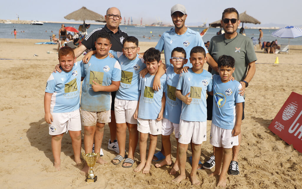 Group of young soccer players in light blue uniforms pose on a sandy beach with two coaches and a trophy in the foreground.