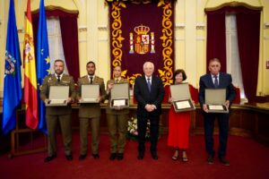 Six people in formal attire hold award plaques during a ceremony in a decorated hall with flags and a regal banner behind them.”, “Military officers and civilians pose with certificates in front of an ornate chamber crest.”, “Officials receive awards in a grand room with flags and a central emblem.”, “Center man in a suit stands with award recipients displaying plaques.”, “Ceremony photo: recipients in uniforms and formal wear display plaques in a ceremonial hall.”