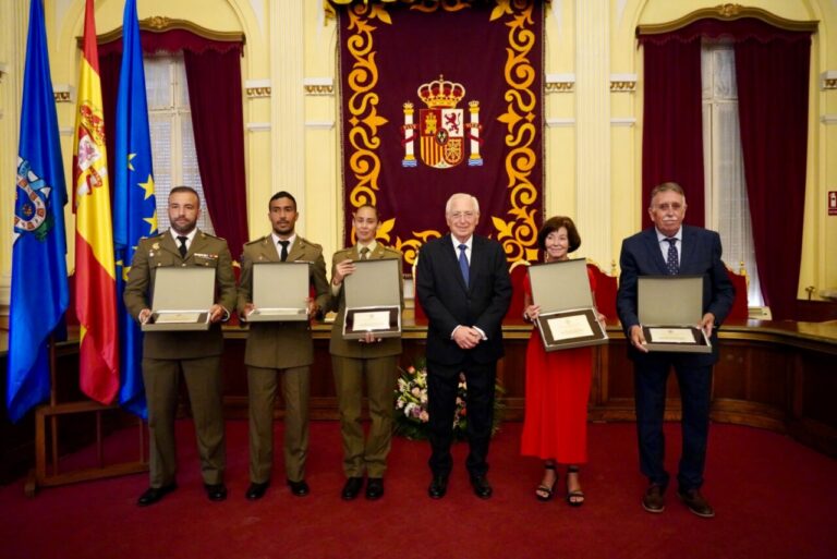 Six people in formal attire hold award plaques during a ceremony in a decorated hall with flags and a regal banner behind them.”, “Military officers and civilians pose with certificates in front of an ornate chamber crest.”, “Officials receive awards in a grand room with flags and a central emblem.”, “Center man in a suit stands with award recipients displaying plaques.”, “Ceremony photo: recipients in uniforms and formal wear display plaques in a ceremonial hall.”