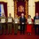 Six people in formal attire hold award plaques during a ceremony in a decorated hall with flags and a regal banner behind them.”, “Military officers and civilians pose with certificates in front of an ornate chamber crest.”, “Officials receive awards in a grand room with flags and a central emblem.”, “Center man in a suit stands with award recipients displaying plaques.”, “Ceremony photo: recipients in uniforms and formal wear display plaques in a ceremonial hall.”