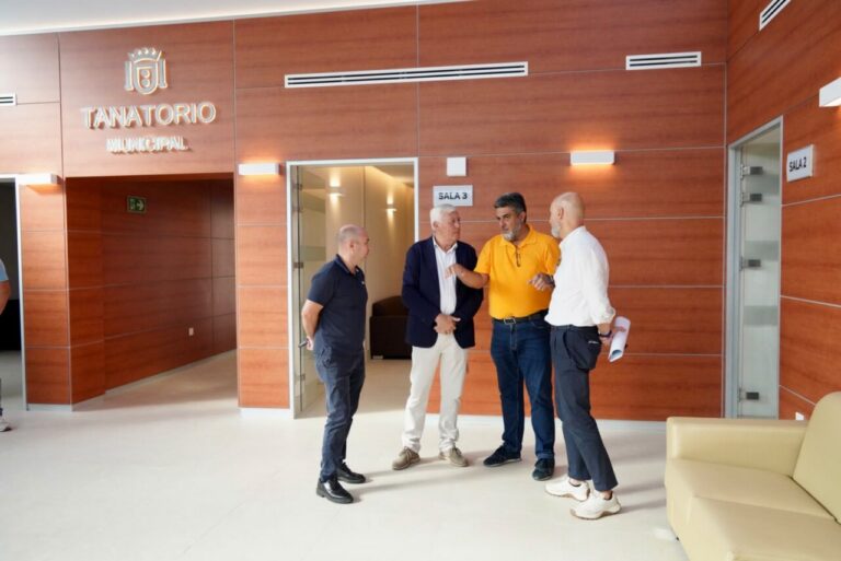 Group of four men standing and talking in a wood-paneled municipal lobby marked 'Tanatorio Municipal' near a doorway labeled Sala 3.