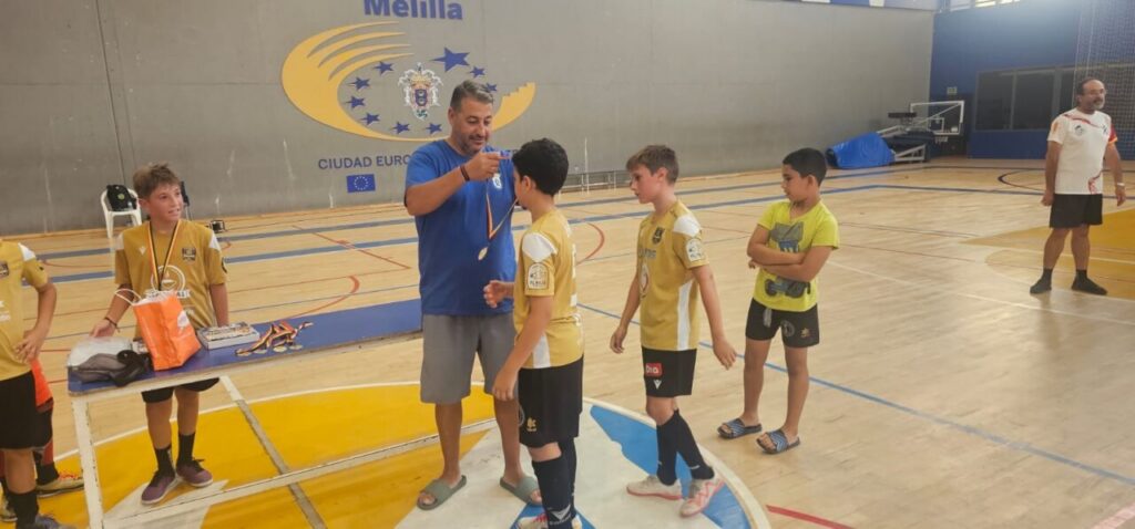 Coach awards medals to a boy during a youth sports ceremony in a gym; teammates in yellow uniforms stand in line nearby while a banner with Melilla is on the wall.]','