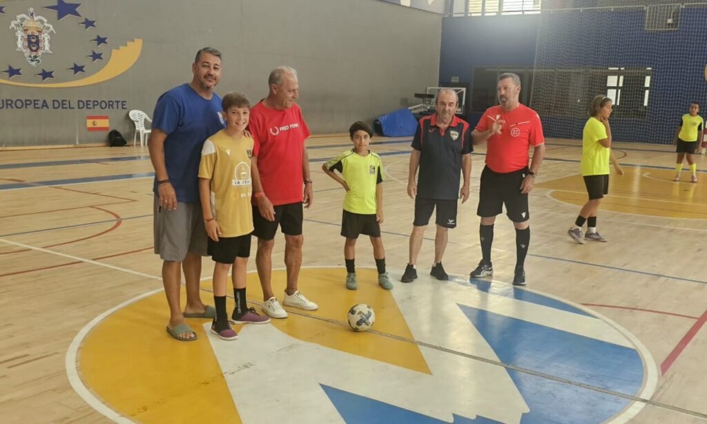 Group of men and two boys posing for a photo on an indoor futsal court, with a soccer ball at their feet and players in yellow shirts in the background.